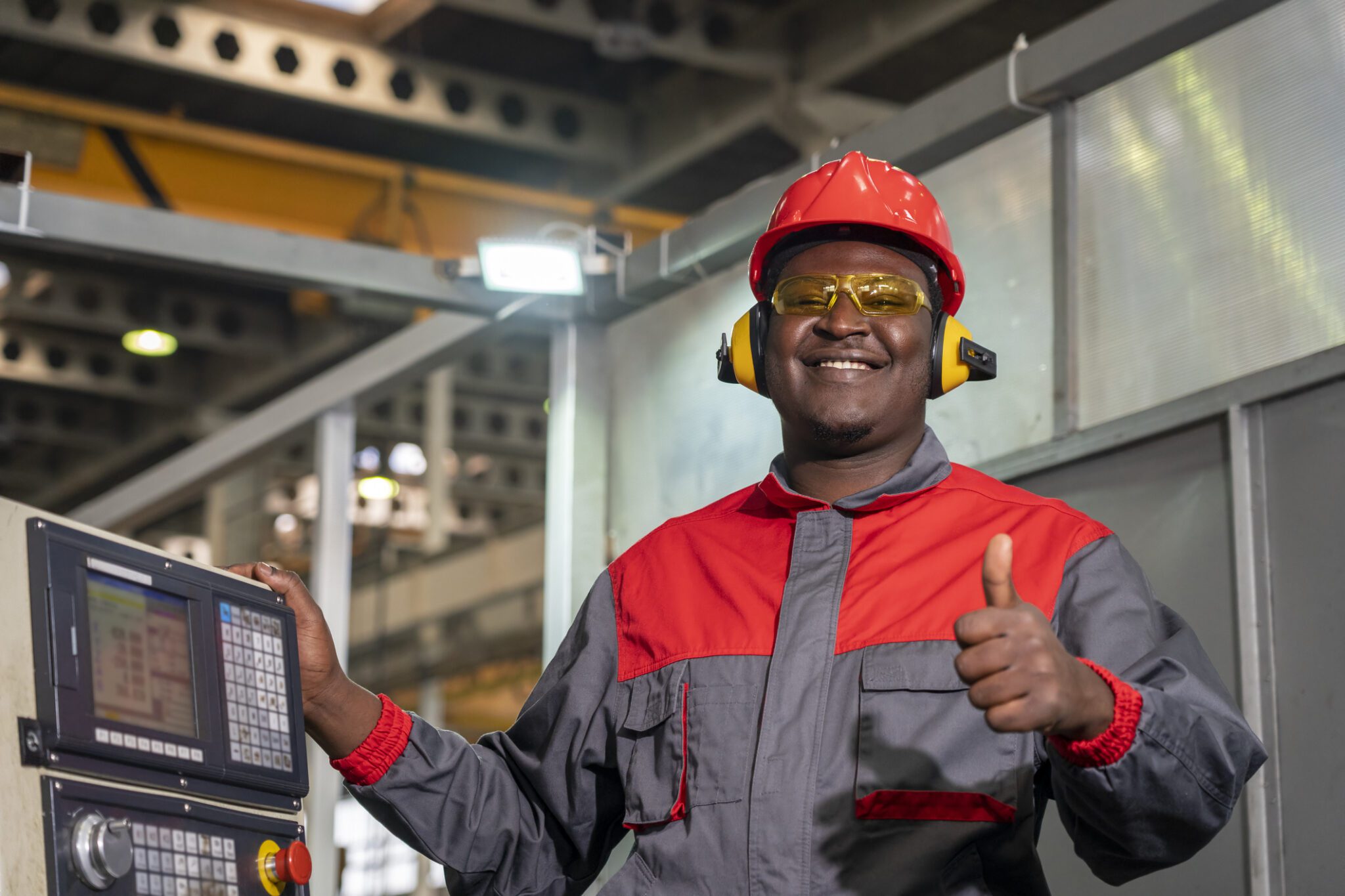 Portrait Of Black Machine Operator In Red Helmet, Safety Goggles, Hearing Protectors And Work Uniform.
